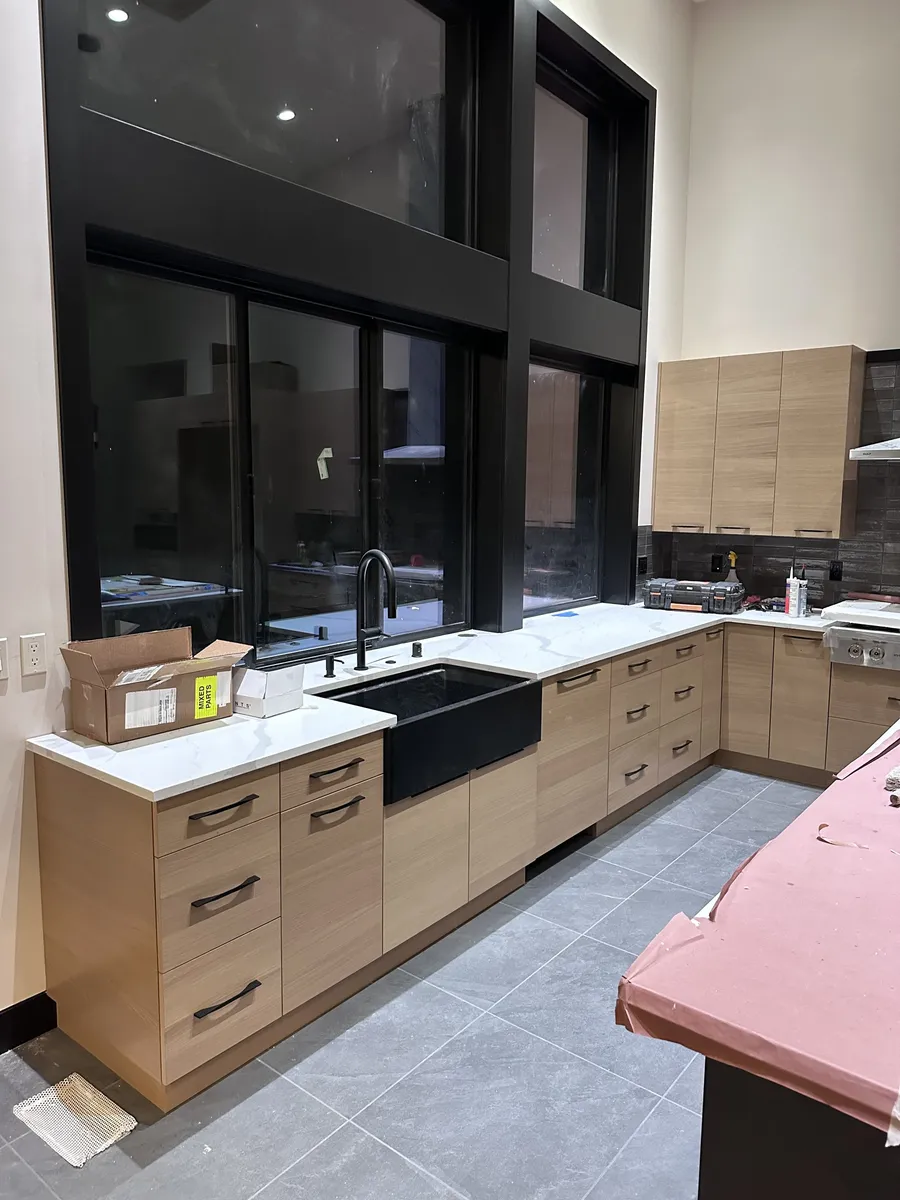 Black farmhouse sink set into light oak base cabinets under tall black windows
